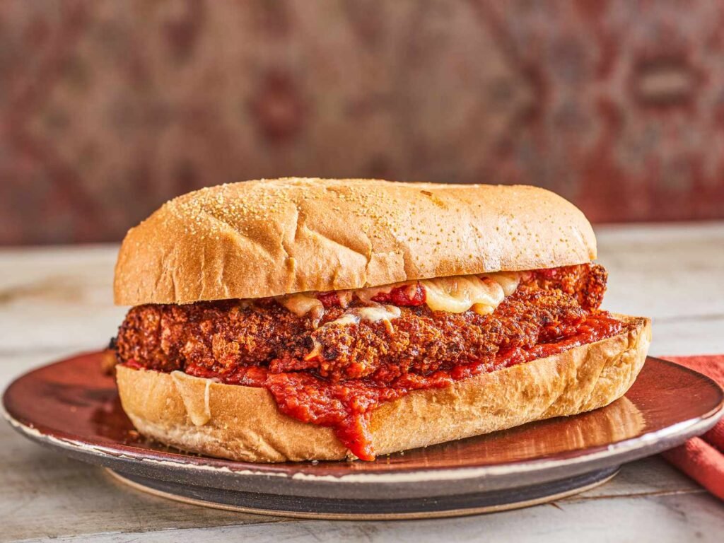 A hearty sandwich on a crusty roll, packed with crisped chicken cutlet, red marinara sauce, and melted cheese, sliced to reveal its juicy interior—a party‑size version from Serious Eats