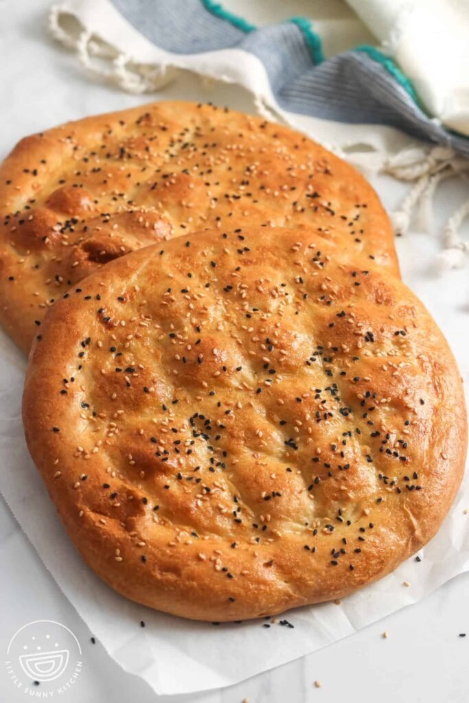 A close-up of a single round pide topped with sesame and nigella seeds, showcasing its shiny, egg-washed crust and the pronounced woven indentations made by finger‑pressing.