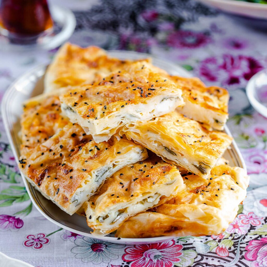 Rectangular tray-baked Börek with crisp, lightly browned top, cut into squares revealing layers of filo pastry and cheese, set on a plate on a floral tablecloth.