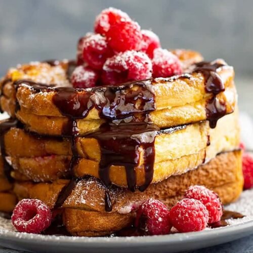 A towering stack of brioche French toast filled with raspberry cheesecake, drizzled with chocolate sauce, and topped with fresh raspberries.