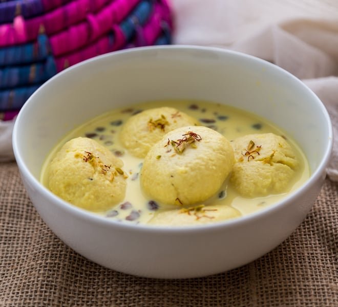 White paneer discs in a bowl of creamy saffron‑infused milk, garnished with chopped pistachios.